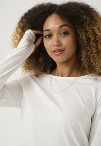 Langärmliges, weißes Oberteil mit glatter Textur. Goldene Halskette mit kleinen, runden Akzenten. Modell mit lockigem Haar, das ihr Haar berührt.