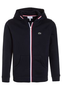 Sudadera con capucha azul marino con un acento en rojo y blanco a rayas, bolsillos tipo canguro y un logo bordado en verde en el lado izquierdo del pecho. Tejido texturizado.