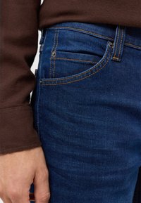 Ciemnoniebieskie dżinsy o widocznym szwie, pięciokomorowym kroju i slim fit. Częściowo widoczny brązowy rękaw swetra w tle.