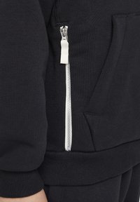 Felpa nera con zip caratterizzata da un tessuto texturizzato, polsini a coste e una zip bianca prominente sulla tasca laterale.