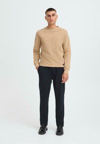 Beige neulepusero, jossa on pyöreä kaula-aukko, yhdistettynä mustiin kapeisiin housuihin ja mustiin kenkiin. Malleilla on yksinkertainen tausta.