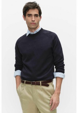 Mężczyzna ubrany w granatowy sweter na jasnoniebieskiej koszuli w paski z podwiniętymi rękawami, beżowe spodnie i brązowy pleciony pasek na tle białego tła.