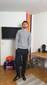 Pilka tekstūruota megztinis, tamsiai mėlynos aptemptos kelnės ir juodi sportbačiai su baltais dryžiais. Vidinė aplinka su televizoriumi ir baldais.