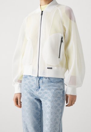 Person iført en hvid net-lynlåstrøje med en forreste brystlomme og lyseblå mønstrede jeans.