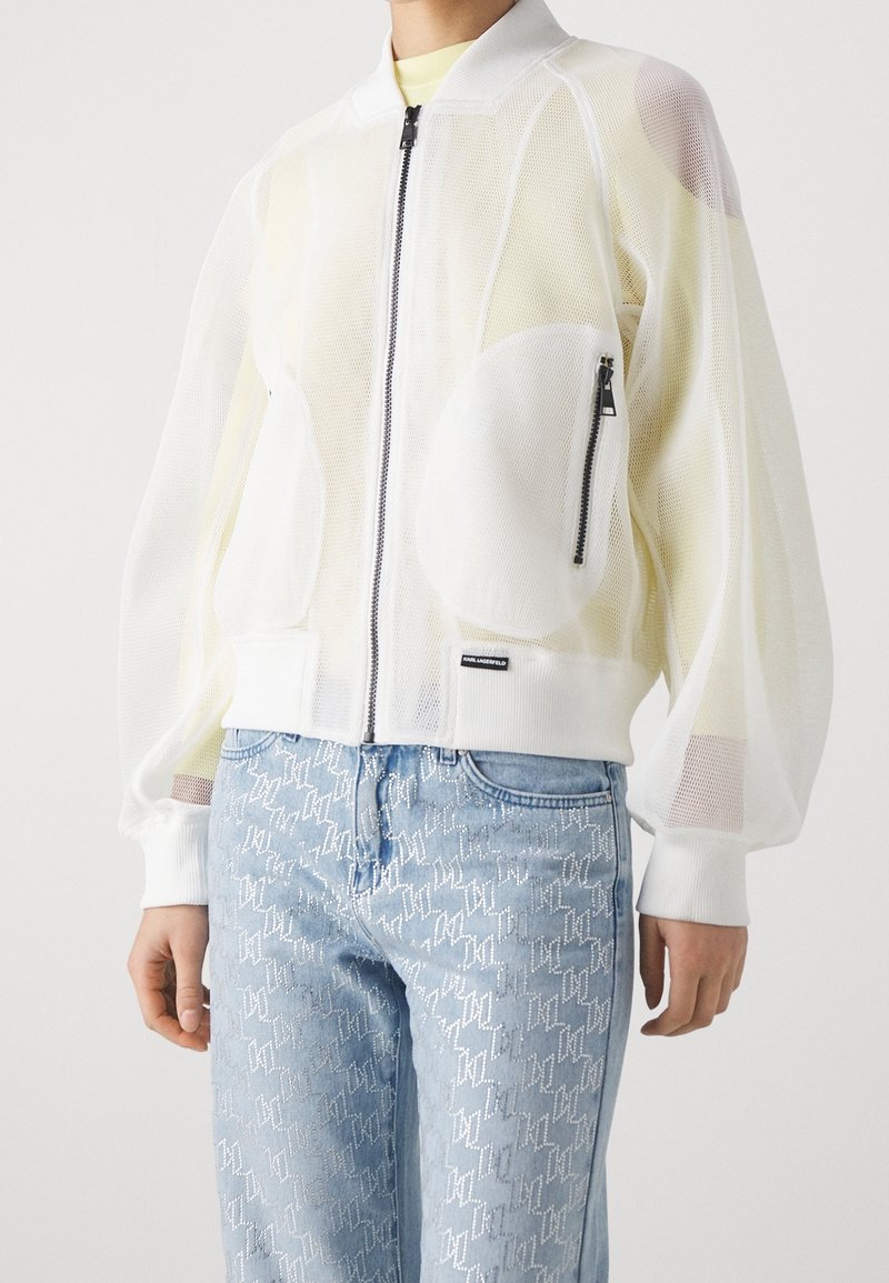 Personne portant une veste blanche en maille zippée avec une poche poitrine avant et un jean bleu clair à motifs.
