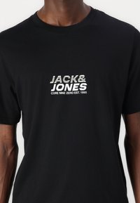 Camiseta de algodón negra con un estampado gráfico blanco que incluye "JACK&JONES" y "CORE NINE ZERO EST. 1990" en la parte delantera. Mangas cortas.