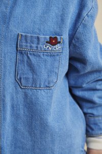 Lyseblå denimskjorte med lange ermer, som har en brystlomme og et brodert design av en rød hatt, med en teksturert stofffinish.