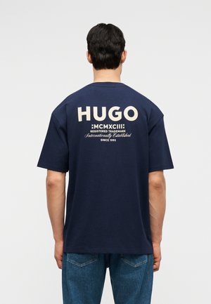 Uomo che indossa una T-shirt blu navy con grande scritta beige sul retro: "HUGO MCMXCIII Marchio Registrato Internazionalmente Stabilito dal 1993".