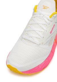 Biele tenisky Reebok s ružovou a žltou podrážkou, bielymi šnúrkami, sieťovaným zvrškom a oranžovým logom Reebok na jazyku a boku.