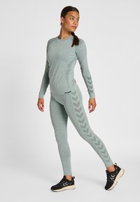 Leichtgewichtiges, strukturiertes grünes Sportset mit langärmligem Oberteil in figurbetonter Passform und Leggings mit Chevron-Akzenten, kombiniert mit schwarzen Turnschuhen.