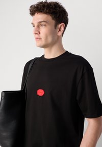 Black cotton t-shirt with a round red logo patch on the chest and a crew neck. The model carries a black leather bag.
