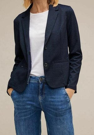 Kobieta w ciemnogranatowej marynarce na białej koszulce i niebieskich dżinsach, z rękami w kieszeniach, na jednolitym beżowym tle.