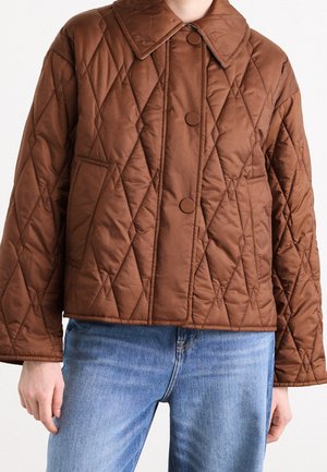 Personne portant une veste matelassée marron avec un grand col et des boutons-pression, associée à un jean en denim bleu.