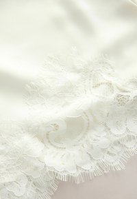 Weißer Stoff mit einem zarten Spitzenbesatz, der filigrane Blumenmuster und leichte Fransen aufweist und eine weiche Textur sowie elegante Details schafft.