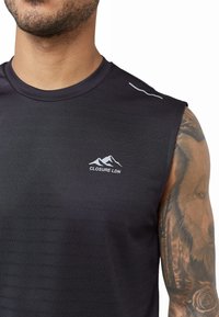 Schwarzes ärmelloses Sportshirt aus strukturiertem Stoff, mit einem weißen "CLOSURE LDN"-Logo und subtilen reflektierenden Akzenten auf den Schultern.