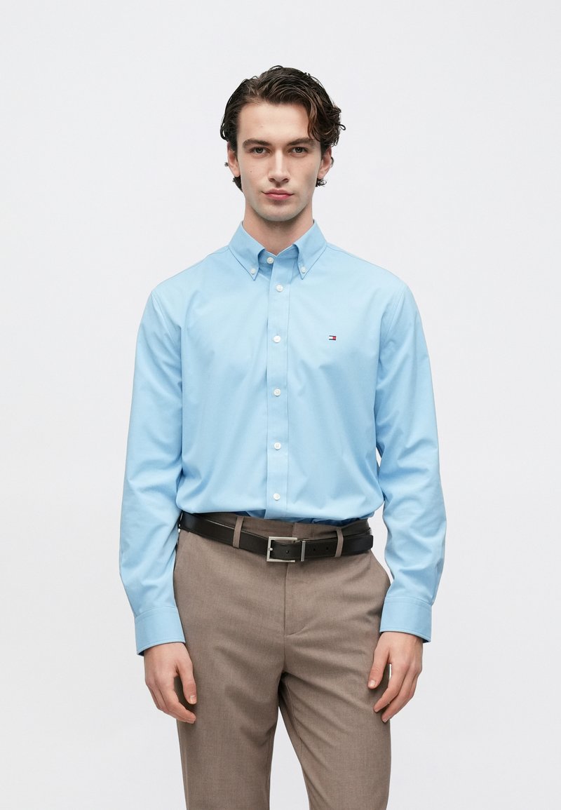 Chemise bleue claire à boutons avec un col, arborant un petit logo sur la poitrine. Associée à un pantalon marron clair et une ceinture noire.