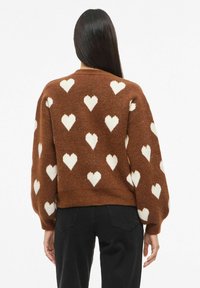 Hnědý svetr s velkými bílými vzory srdcí, dlouhými rukávy a zaobleným výstřihem. Měkká textura, cropovaný střih a žebrovaný lem.