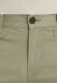 Nahaufnahme von beigen Hosen mit einem braunen Knopf und eingenähtem Reißverschluss, kombiniert mit einem hellbeigen, gerippten Pullover an der Taille.