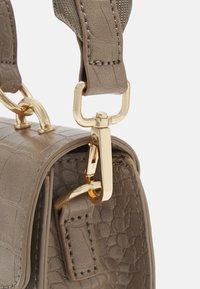 Poignée de sac à main en cuir taupe texturé fixée à un fermoir et un anneau en métal doré avec coutures visibles et détails en gros plan.