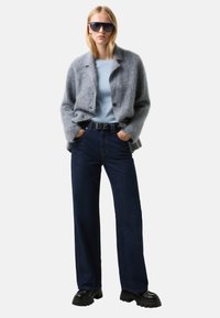Blondynka w oversize'owym, szarym, futrzanym kardiganie, jasnoniebieskiej bluzce, ciemnych szerokich dżinsach, czarnych, masywnych butach i dużych czarnych okularach przeciwsłonecznych, stojąca z rękami w kieszeniach.