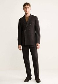 Giacca di abito doppiopetto di colore marrone scuro con tasche laterali, abbinata a pantaloni coordinati e scarpe in suede marroni. Tessuto liscio e vestibilità su misura.