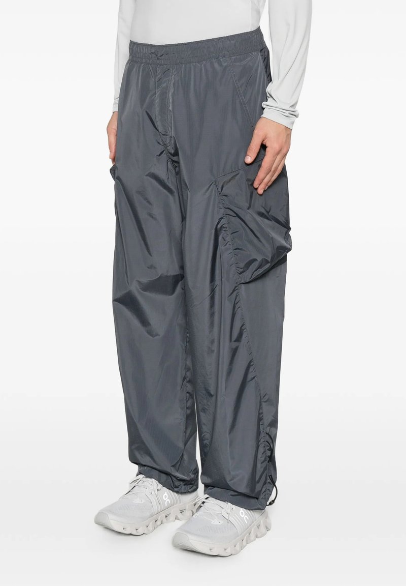 Pantaloni larghi in nylon grigio scuro con elastico in vita e grande tasca laterale, indossati con sneakers bianche e camicia bianca a maniche lunghe.