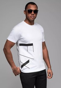 Camiseta blanca con líneas gráficas negras en la parte frontal, cuello redondo y mangas cortas. Combinada con pantalones oscuros y gafas de sol.