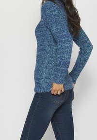 Pull en tricot bleu avec un motif texturé, manches longues et coupe décontractée, port�é avec un jean en denim foncé. Le tissu semble doux et chaud.