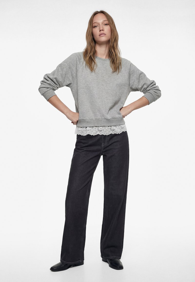 Frau steht mit den Händen in der Hüfte, trägt einen grauen Sweatshirt mit Spitzenbesatz, schwarze weite Hosen und schwarze Schuhe vor weißem Hintergrund.