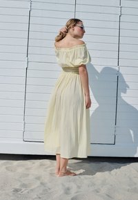 Maxiklänning i gul gingham med barbara axlar, puffiga ärmar och resår i midjan, mot en vit träbakgrund.