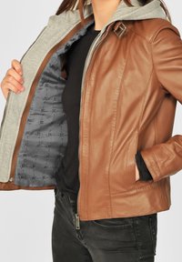 Braune Lederjacke mit grauem Kapuzen, Reißverschluss vorne und Seitentaschen. Innenausstattung mit gemustertem grauem Futter. Glatte Textur.