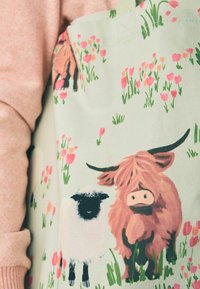 Katoenen schort met een lichtgroene achtergrond, geïllustreerd met schapen en een Hooglandrund te midden van roze bloemen en grasaccenten.