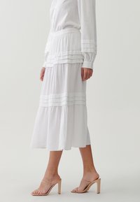 Falda blanca de capas con pliegues horizontales y un corte suelto. Fabricada en un tejido ligero. Lleva unas sandalias de tacón nude.
