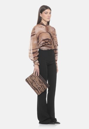 Kobieta ubrana w brązową, wzorzystą, prześwitującą bluzkę oraz czarne spodnie, trzymająca brązową kopertówkę w tygrysie paski.
