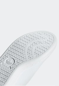 Semelle de chaussure blanche avec un motif hexagonal texturé et des lignes courbes. Présente un logo au talon, des bords lisses et une semelle discrète pour l'adhérence.