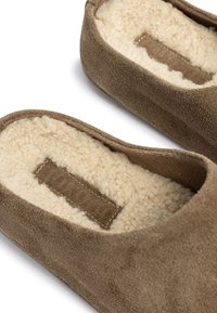 Zapatillas de ante marrón con un suave forro de piel de cordero en color crema, que presentan una etiqueta texturizada en tonos marrones y un diseño sencillo y redondeado.