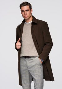 Ruskea villainen takki harmaan neuleen päällä, yhdistetty harmaan ja kerman sävyisiin ruutuhousuihin. Ranteessa näkyy kello, sileät tekstuurit kauttaaltaan.