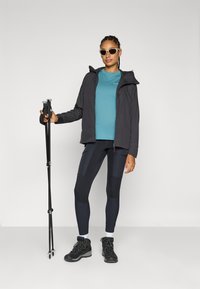 Wanderoutfit mit einer schwarzen Jacke, einem türkisfarbenen Shirt, navyblauen Leggings mit unterschiedlichen Texturen und schwarzen Stiefeln, während Trekkingstöcke gehalten werden.