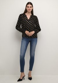Blusa nera con motivo a foglia d'oro; maniche lunghe con polsini arricciati, scollatura a V. Abbinata a jeans skinny blu e tacchi a punta neri.