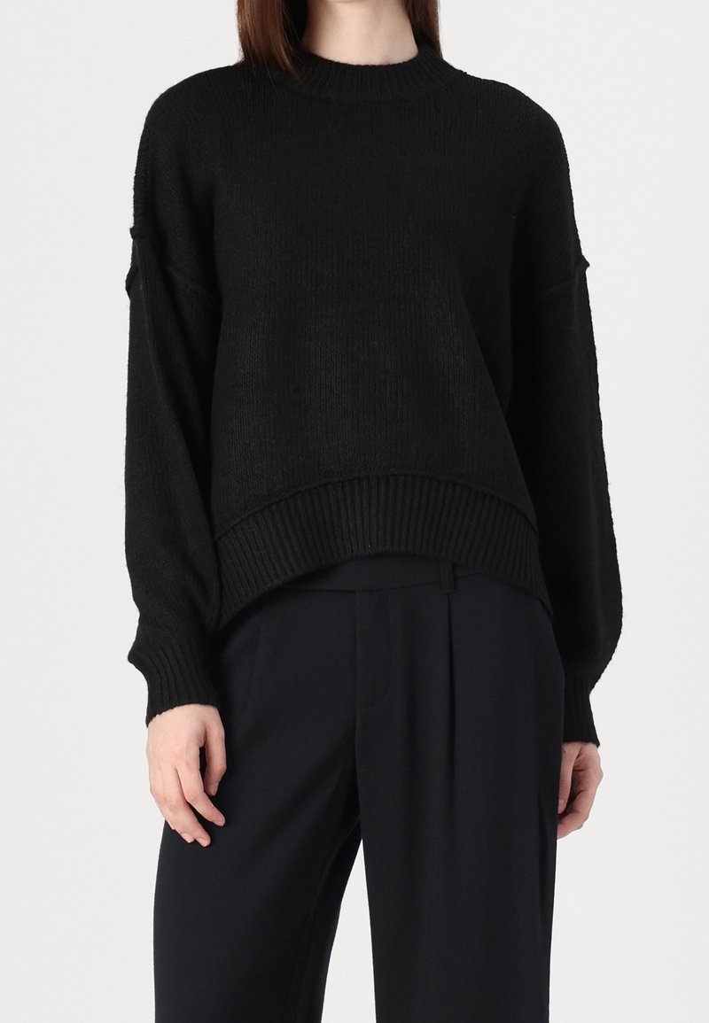 Schwarzer gestrickter Pullover mit strukturierter Oberfläche, Rundhalsausschnitt, überschnittenen Schultern und geripptem Saum. Kombiniert mit dunkelgrauen Anzughosen.