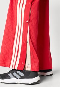 Czerwone dresowe spodnie z siateczki z białymi paskami i guzikami po bokach, zestawione z czarnymi butami sportowymi z szarymi akcentami.