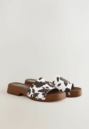 Sandale cu imprimeu de vacă, cu o curea lată, toc din lemn și nituri decorative aurii pe laturi. Talpă plată cu o suprafață texturată.