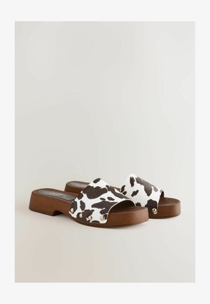 Koeienprint slippers met een brede band, houten blokhak en gouden accentstuds aan de zijkanten. Vlakke zool met een textuur oppervlak.