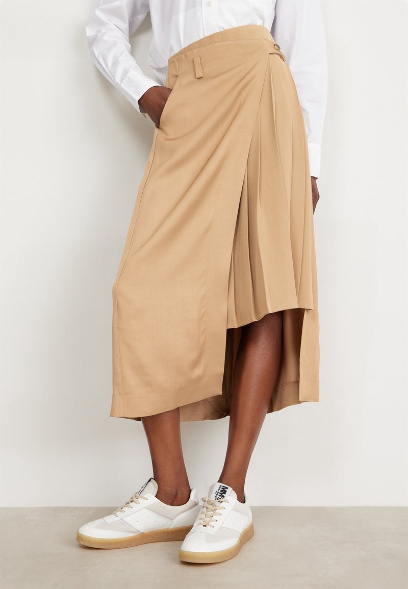 Jupe beige midi avec un ourlet asymétrique, un design plissé, des poches latérales et une ceinture ajustée, associée à des baskets blanches.