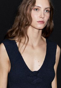 Námořnický modrý pletený top s hlubokým výstřihem do V a květinovým akcentem uprostřed. Textura je žebrovaná a těsně přiléhá.