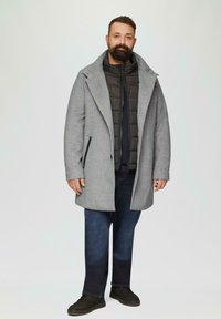 Grijze wollen overjas met een brede kraag over een zwarte pufferjack. Donkerdenim jeans en zwarte suède schoenen maken de look compleet.