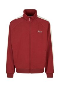 Červená bunda na zips s vysokým golierom, biele bočné pruhy na rukávoch, predné vrecká a logo "Kani" na ľavej hrudi.