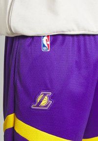 Lila basketshorts med gula ränder, med en broderad Lakers-logga och en NBA-tag. Tillverkade av strukturerat meshmaterial.