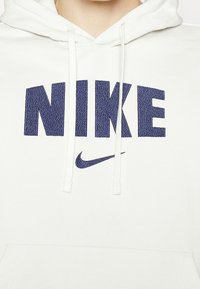 Vit hoodie med en marinblå broderad "NIKE"-logotyp och swoosh. Har dragsnören och en texturerad tygfinish.