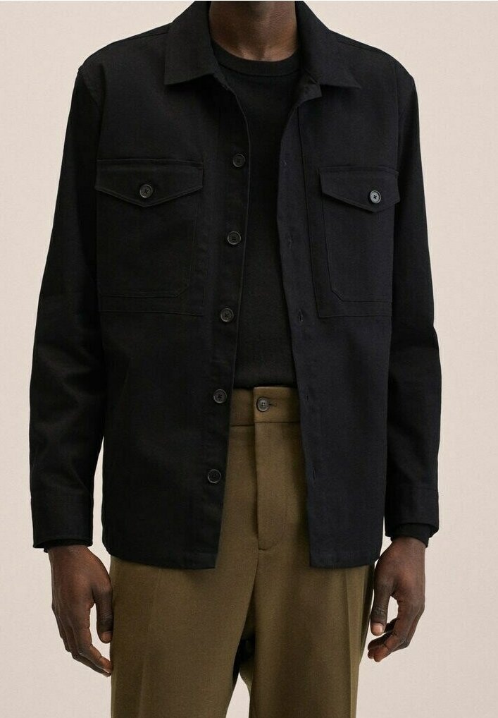 Personne portant une veste noire boutonnée sur une chemise noire avec un pantalon taille haute vert olive, les mains détendues le long du corps.